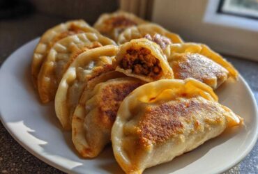 A plate of golden brown, pan-fried dumplings, with one dumpling cut open showing the savory filling.