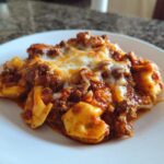 A close-up serving of rich Beef Enchilada Tortellini topped with melted white cheese on a white plate.