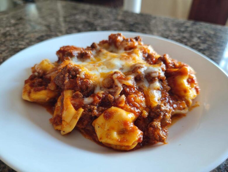 A close-up serving of rich Beef Enchilada Tortellini topped with melted white cheese on a white plate.