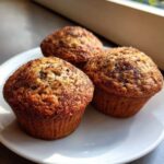 Three freshly baked Brown Sugar Banana Muffins with a sugary, spiced crust, resting on a white plate.