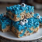 A stack of four bright blue Cookie Monster Rice Krispy Treat Bars topped with candy eyeballs on a white plate.