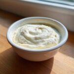 A small white bowl filled with thick, rich Creamy White Garlic Pizza Sauce, showing swirls and specks of herbs.