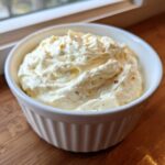 A white ramekin filled with thick, rich Creamy White Garlic Pizza Sauce, resting on a wooden surface near a window.