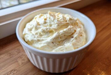 A white ramekin filled with thick, rich Creamy White Garlic Pizza Sauce, resting on a wooden surface near a window.