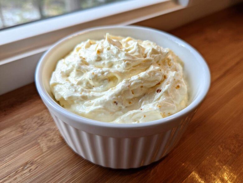 A white ramekin filled with thick, rich Creamy White Garlic Pizza Sauce, resting on a wooden surface near a window.