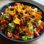 A close-up view of a hearty Doritos Taco Salad featuring seasoned ground beef, shredded cheese, olives, tomatoes, and tortilla chips over lettuce.