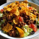 A vibrant bowl of Doritos Taco Salad featuring seasoned ground beef, lettuce, tomatoes, black olives, and crushed Doritos chips.