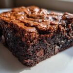 A close-up, macro shot of a rich, fudgy brownie square showing its shiny, crackled top and visible chocolate chips.