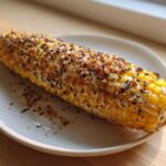 A close-up of a cooked ear of Garlic Parmesan Corn on the Cob generously topped with cheese, spices, and black sesame seeds.