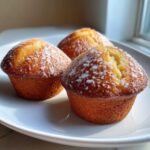Three golden brown Keto Lemon Muffins topped with a light white glaze, resting on a white plate near a window.