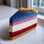 A perfect slice of layered Red White and Blue Cheesecake showing blue, pink, white, and red layers on a graham cracker crust.