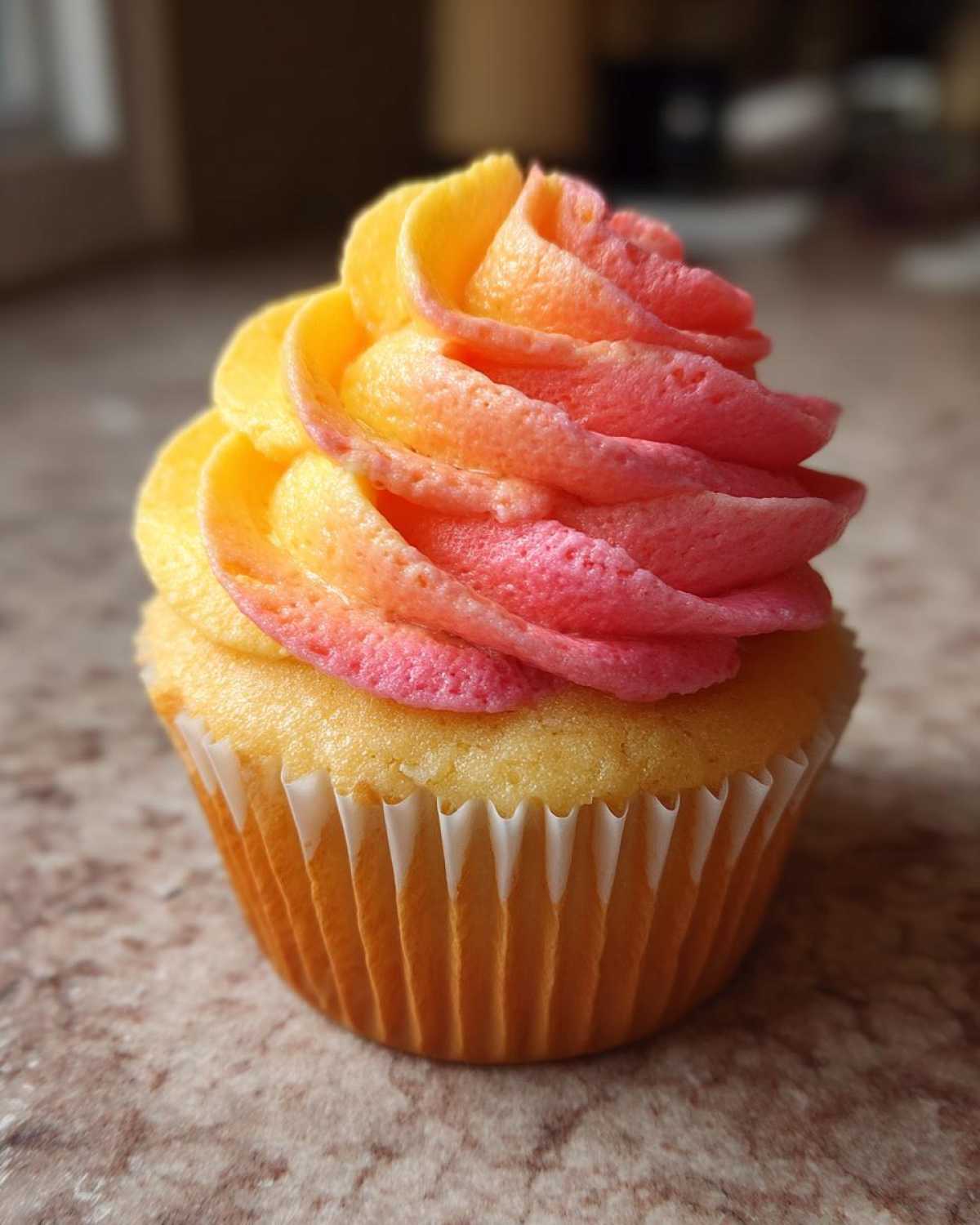 A single cupcake topped with vibrant, swirled frosting featuring yellow, orange, and pink colors, resembling a sunset, for Mango Strawberry Sunset Cupcakes.