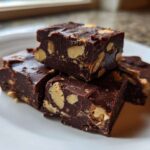 Close-up of several squares of rich, dark Microwave Snickers Fudge loaded with peanuts, stacked on a white plate.