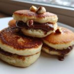 Close-up of stacked Mini Bananas Pancakes with Nutella filling, topped with hazelnuts, served on a white plate.