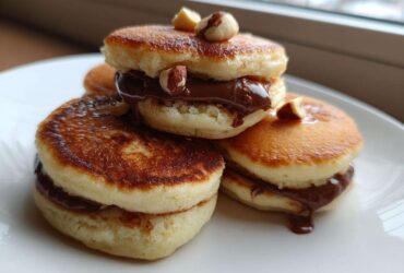 Close-up of stacked Mini Bananas Pancakes with Nutella filling, topped with hazelnuts, served on a white plate.