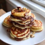 A stack of golden Mini Bananas Pancakes layered with melted Nutella and topped with hazelnuts.