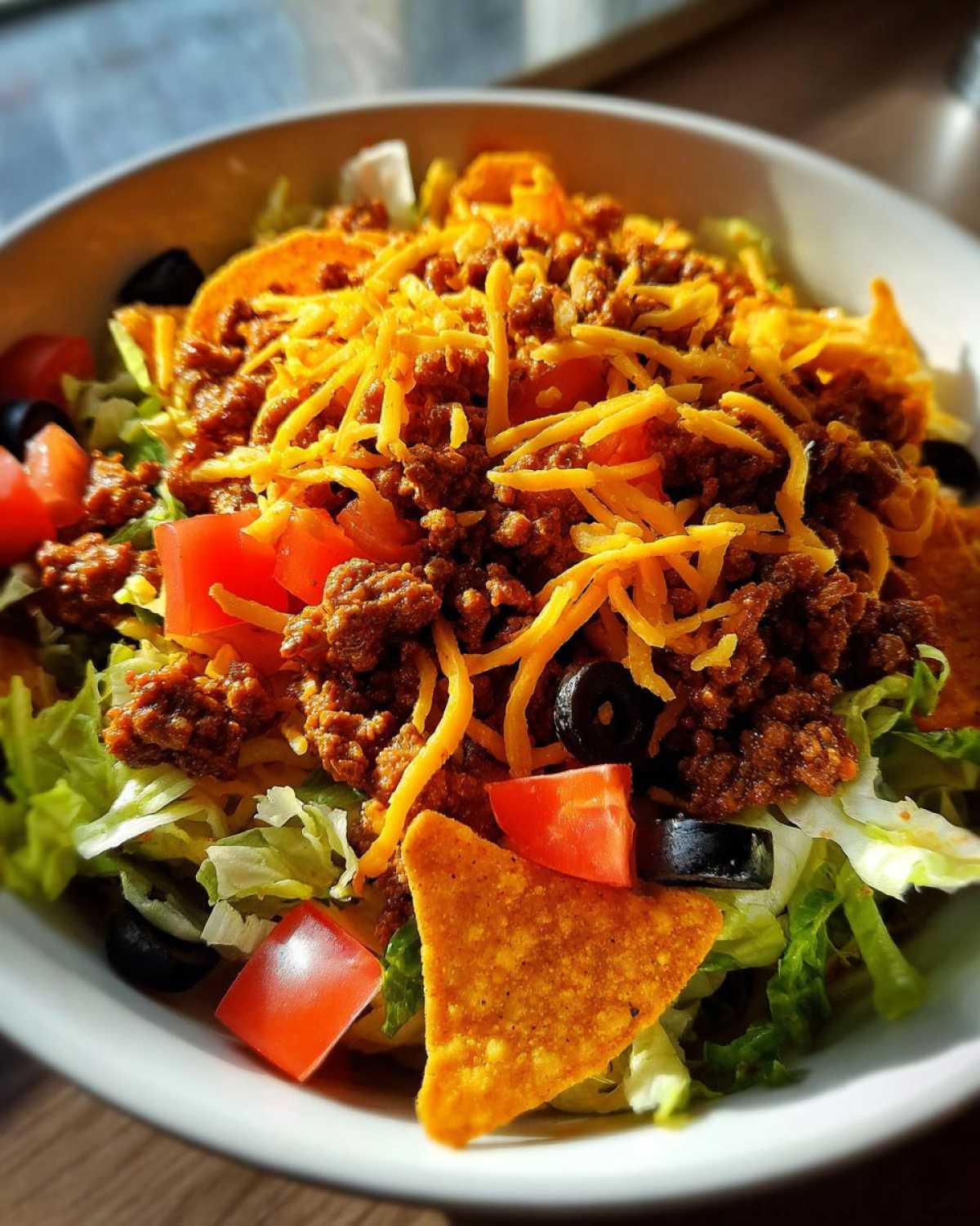 A close-up view of a vibrant Doritos Taco Salad bowl topped with seasoned ground beef, shredded cheese, tomatoes, and black olives.