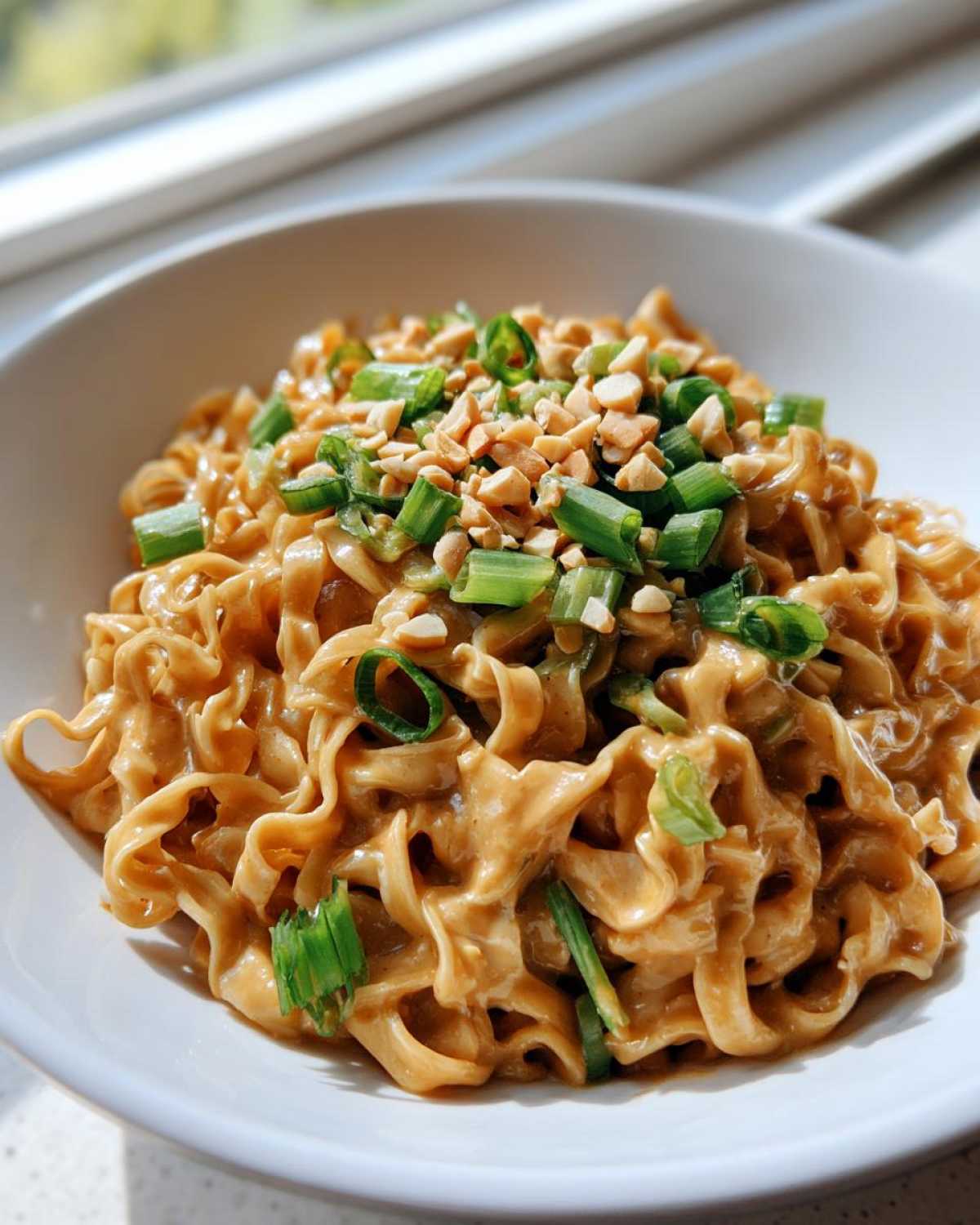 A white bowl filled with creamy Saucy Ramen Noodles, topped with chopped green onions and crushed peanuts.