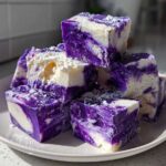 A stack of vibrant purple and white marbled Blueberry Fudge squares dusted with powdered sugar on a light plate.