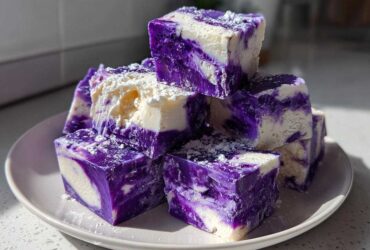 A stack of vibrant purple and white marbled Blueberry Fudge squares dusted with powdered sugar on a light plate.